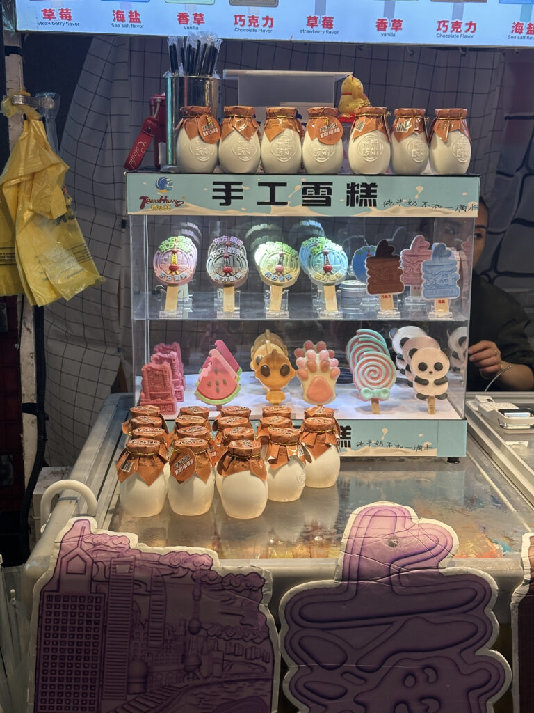 The image shows a display of various ice cream and dessert items in a shop. On the top shelf, there are several jars with brown foil lids, possibly containing a type of dessert or yogurt. Below, there are colorful ice cream bars in various shapes, including a watermelon slice, a paw print, a swirl lollipop, and a panda face. The display is labeled with Chinese characters, and there are signs above indicating different flavors such as strawberry, sea salt, vanilla, and chocolate. The setup is bright and colorful, attracting attention to the variety of treats available.
