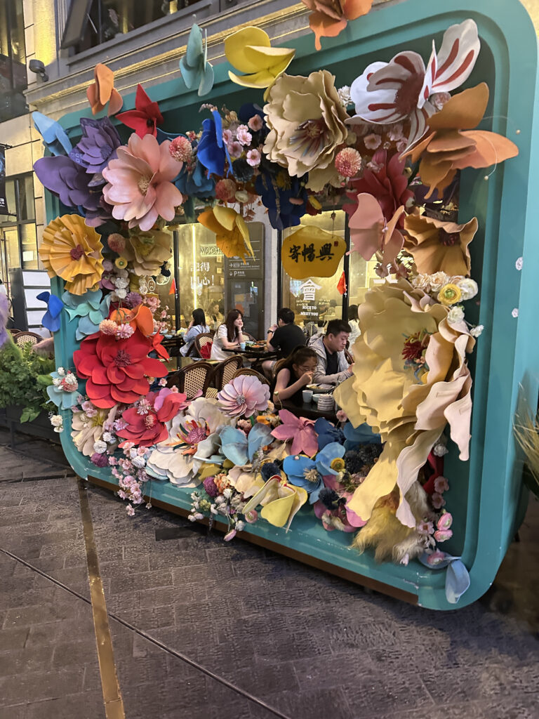 The image shows a decorative frame adorned with large, colorful paper flowers in various shapes and colors, including red, pink, blue, yellow, and orange. Inside the frame, there are people sitting at a table in an outdoor dining area, with a restaurant visible in the background. The setting appears to be lively and vibrant, with a mix of natural and artificial lighting.