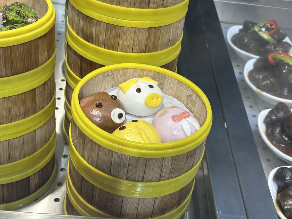 A bamboo steamer basket containing four colorful, animal-shaped buns. Each bun is designed to resemble a different animal, with distinct features like ears and eyes. The basket is part of a stack with yellow rims, and there are other dishes visible in the background.