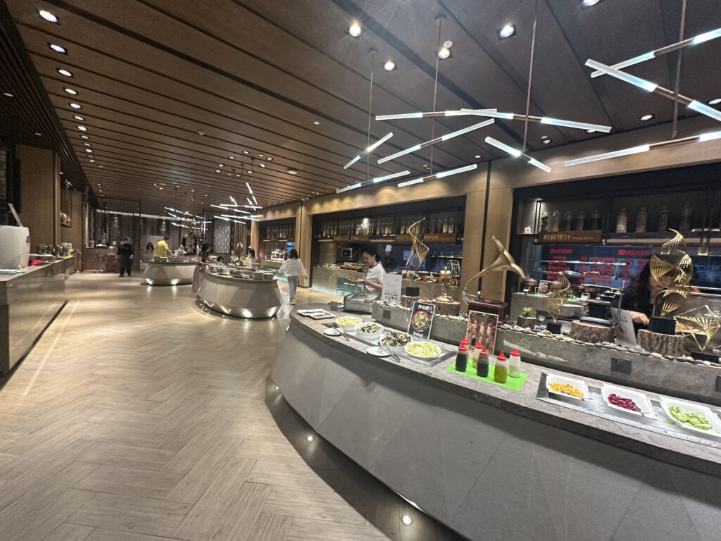 The image shows a modern buffet restaurant with a sleek, contemporary design. The interior features a long counter with various food items displayed, including plates of vegetables and sauces. The ceiling has stylish lighting fixtures, and there are decorative elements on the counter. Several people are visible in the background, serving or selecting food. The overall ambiance is upscale and well-lit.