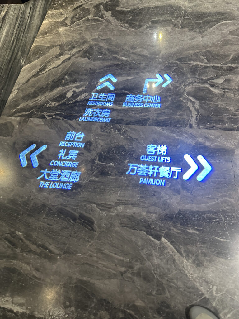 The image shows a marble floor with illuminated directional signs in both Chinese and English. The signs point to various facilities: "Restrooms," "Business Center," "Laundromat," "Reception," "Concierge," "The Lounge," "Guest Lifts," and "Pavilion." The text is glowing in blue, and arrows indicate the directions to each location.