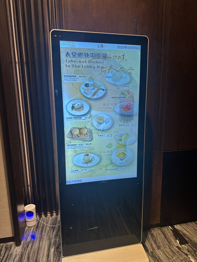 The image shows a digital display board in a hotel lobby, featuring a menu for take-out dishes. The menu includes various items such as mousse cakes, Swiss rolls, yogurt, berry lemon tea, pineapple buns, mango pudding, and more. The display is titled "Take-out Dishes In The Lobby Bar" and includes both English and Chinese text. The date on the screen is June 9, 2025, and the location is indicated as Shanghai.