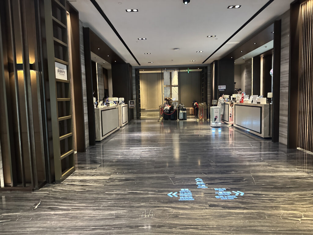 The image shows a modern hotel lobby with a sleek, polished floor and contemporary design elements. There are two reception desks on either side, each staffed by people. The lighting is soft and warm, with ceiling lights providing illumination. In the center, there is a display of various items, possibly promotional or decorative. The overall atmosphere is clean and professional.