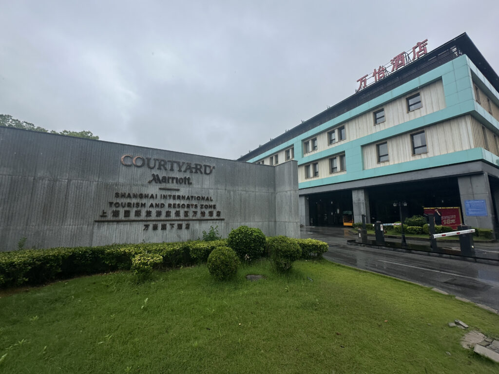 The image shows the exterior of a Courtyard by Marriott hotel located in the Shanghai International Tourism and Resorts Zone. The building has a modern design with a light blue and white facade. In the foreground, there is a sign with the hotel's name and location details, surrounded by neatly trimmed bushes and a grassy area. The sky is overcast, suggesting a cloudy day.
