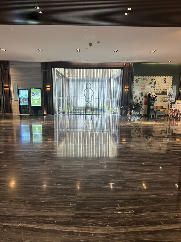 The image shows a modern indoor space with a polished, reflective floor. In the center, there is a decorative water feature with vertical streams of water and a geometric design on the wall behind it. On the left, there are digital screens displaying information. On the right, there is a display with panda-themed decorations, including a bicycle and a large stamp design on the wall. The ceiling has recessed lighting.