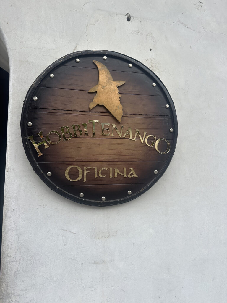 A round wooden sign mounted on a white wall, featuring the silhouette of a wizard's hat and face in gold. The text on the sign reads "Hobbitenango Oficina" in a stylized font. The sign is bordered with metal studs.