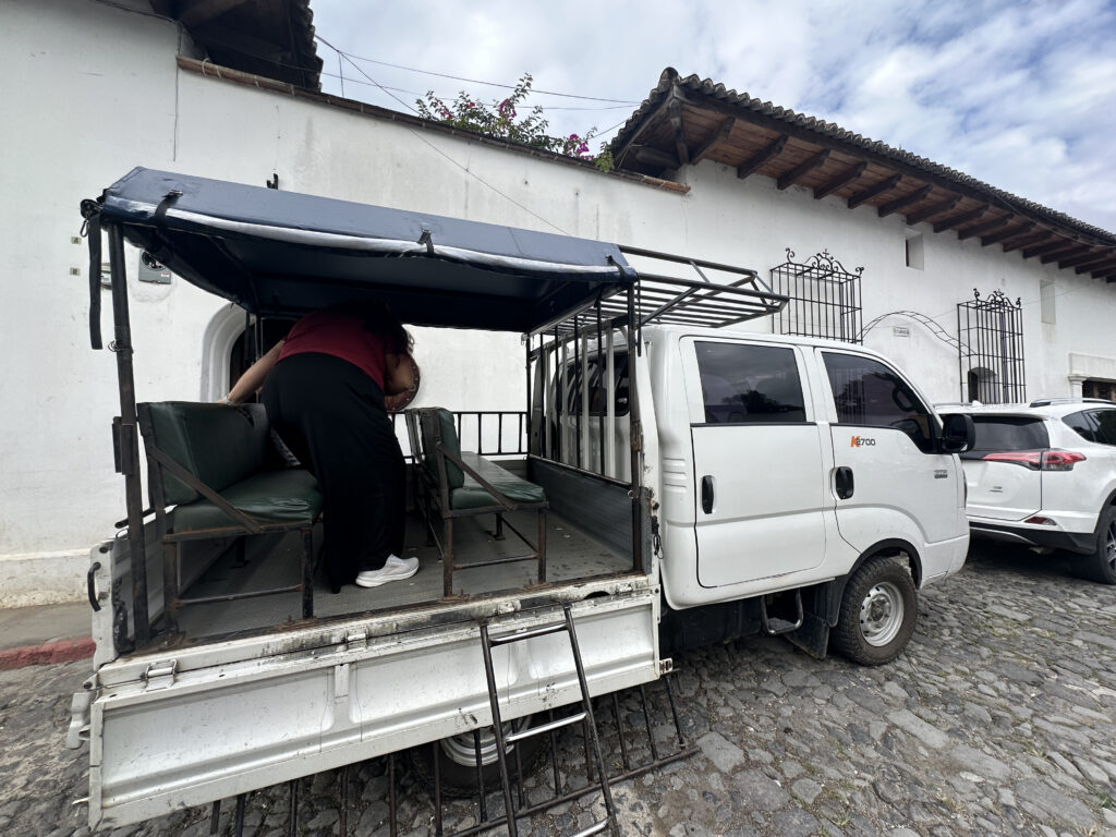 A white pickup truck with an open back is parked on a cobblestone street. The back of the truck has a canopy and bench seating. A person is leaning into the back of the truck. The background features a white building with wooden roof tiles and decorative wrought iron window bars. Another white vehicle is parked nearby.