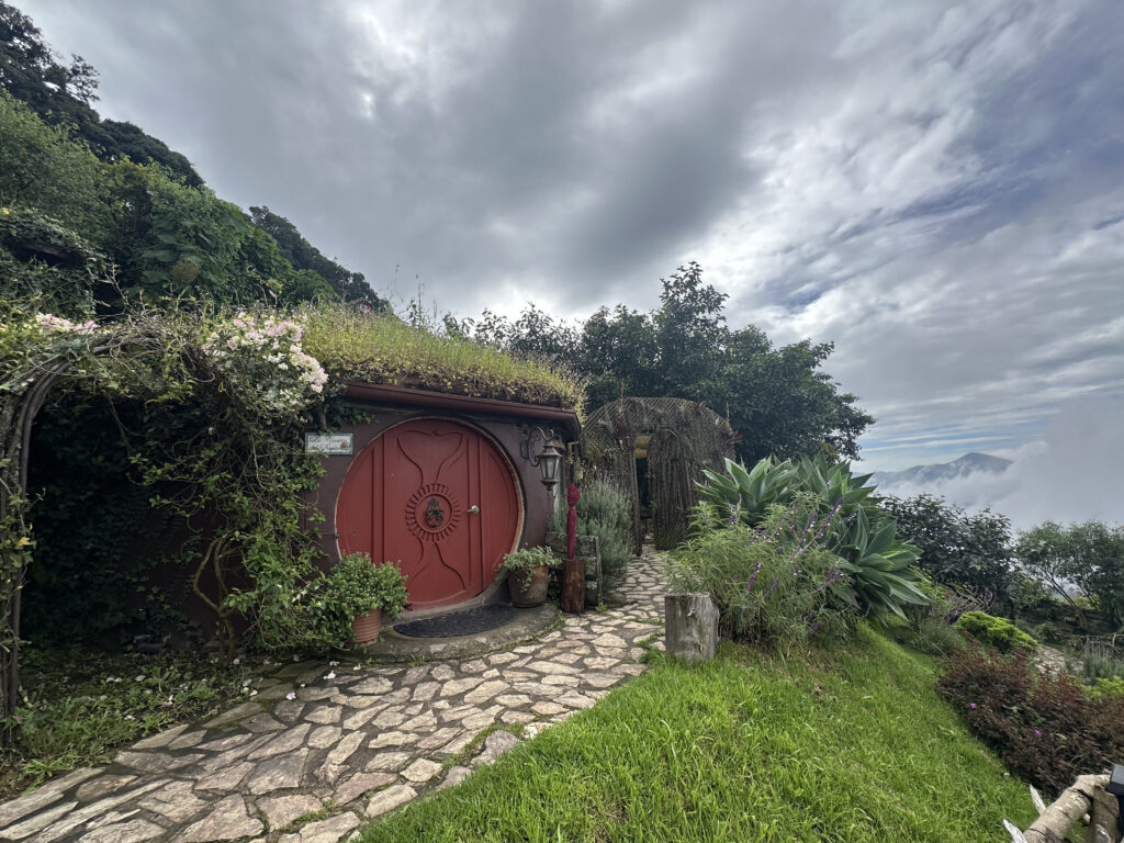 A small, round-door house built into a hillside, resembling a hobbit home. The door is red with a circular design, surrounded by lush greenery and flowers. The roof is covered with grass and plants. A stone path leads to the entrance, and the background features a cloudy sky and distant mountains.