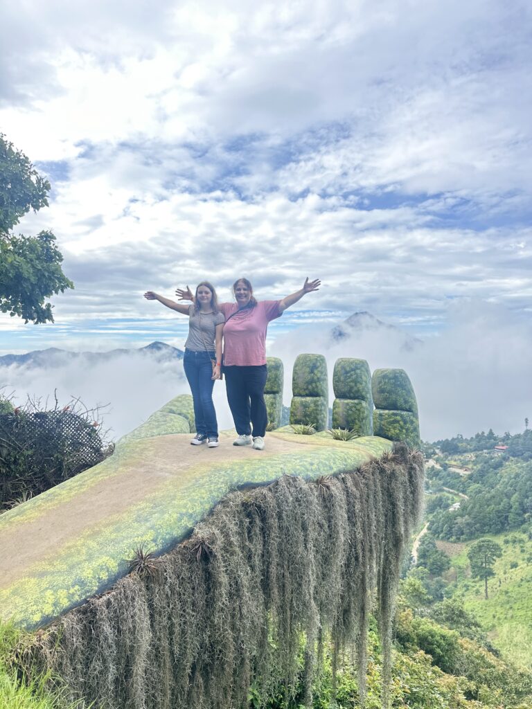 Two people are standing on a large, sculpted hand platform that appears to be part of a scenic overlook. The hand is covered in greenery and moss, with long, hanging plants draping from the edge. They are surrounded by a cloudy sky and a lush, mountainous landscape in the background. Both individuals have their arms outstretched, enjoying the view.