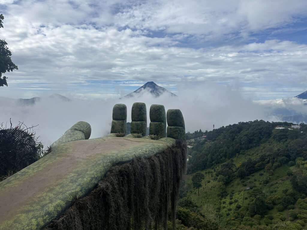 A large sculpture of a hand extends outward, appearing to emerge from the landscape. It is covered in moss or a similar green texture. The background features a scenic view of mountains partially obscured by clouds, with lush greenery in the foreground. The sky is mostly cloudy with patches of blue.