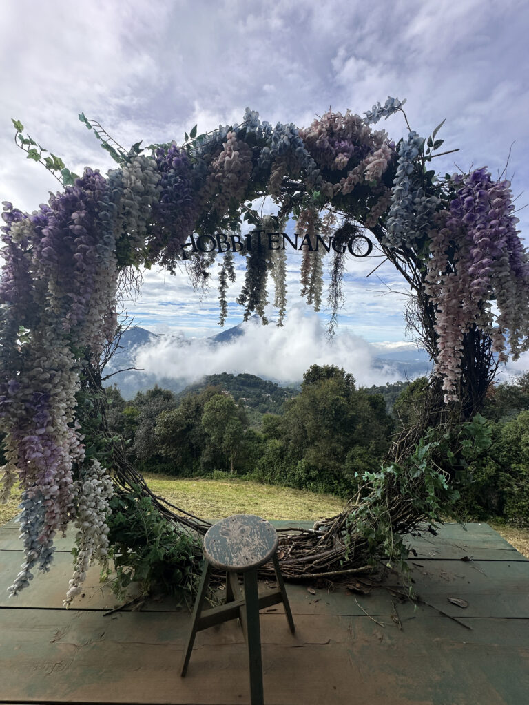 A large circular floral arch made of intertwined branches and adorned with cascading flowers in shades of purple, pink, and white is set against a scenic backdrop of mountains and clouds. A small wooden stool is placed in front of the arch on a wooden platform. The sky is partly cloudy, adding to the serene and picturesque setting.