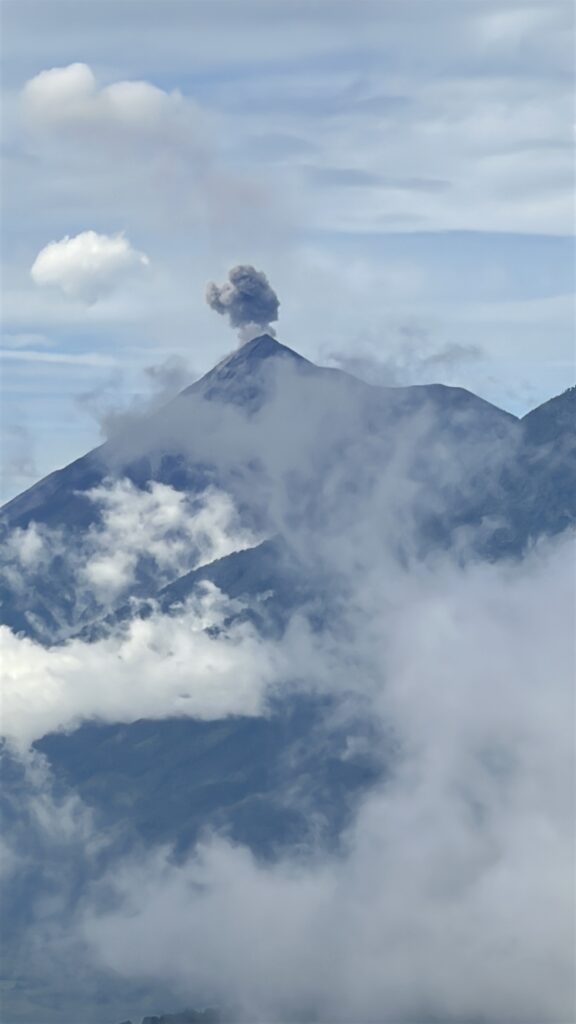A volcanic mountain peak is partially obscured by clouds, with a plume of smoke rising from the summit against a blue sky.