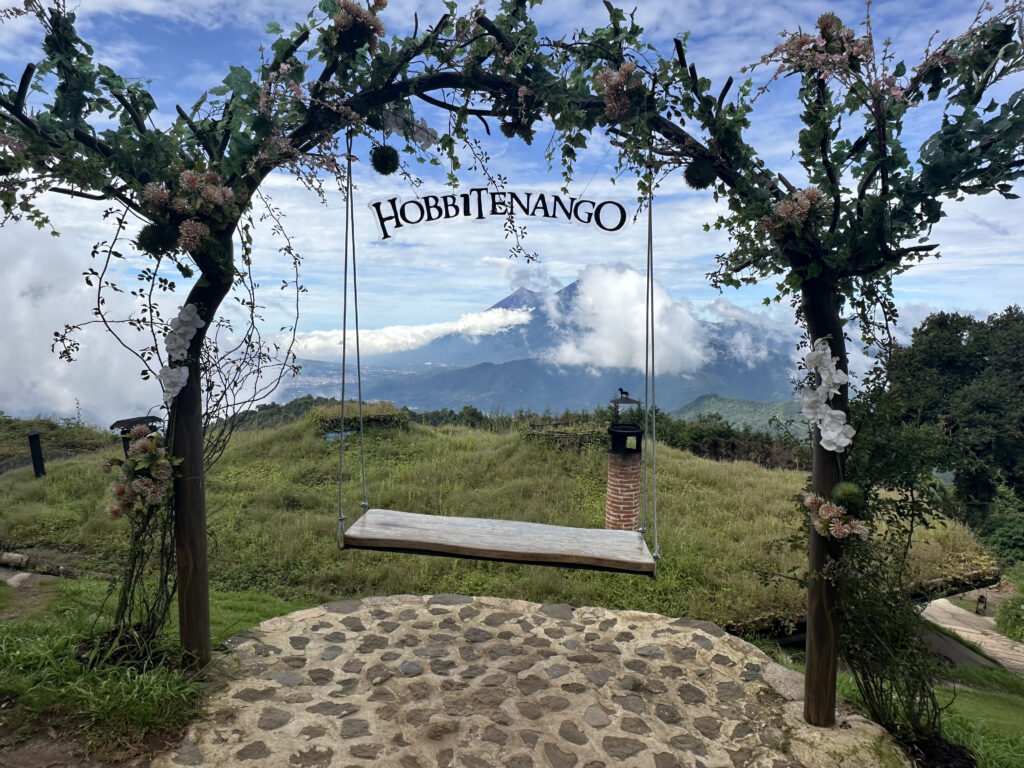 A wooden swing hangs from an archway adorned with green leaves and pink flowers. The archway has the word "Hobbitenango" on it. In the background, there is a scenic view of mountains partially covered by clouds, under a blue sky. The foreground features a stone path and grassy area.