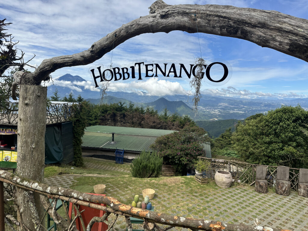A scenic view of a mountainous landscape with a sign reading "Hobbitenango" hanging from a wooden arch. In the foreground, there is a rustic fence and a patio area with wooden chairs and potted plants. The background features lush greenery, a building with a green roof, and distant mountains under a partly cloudy sky.