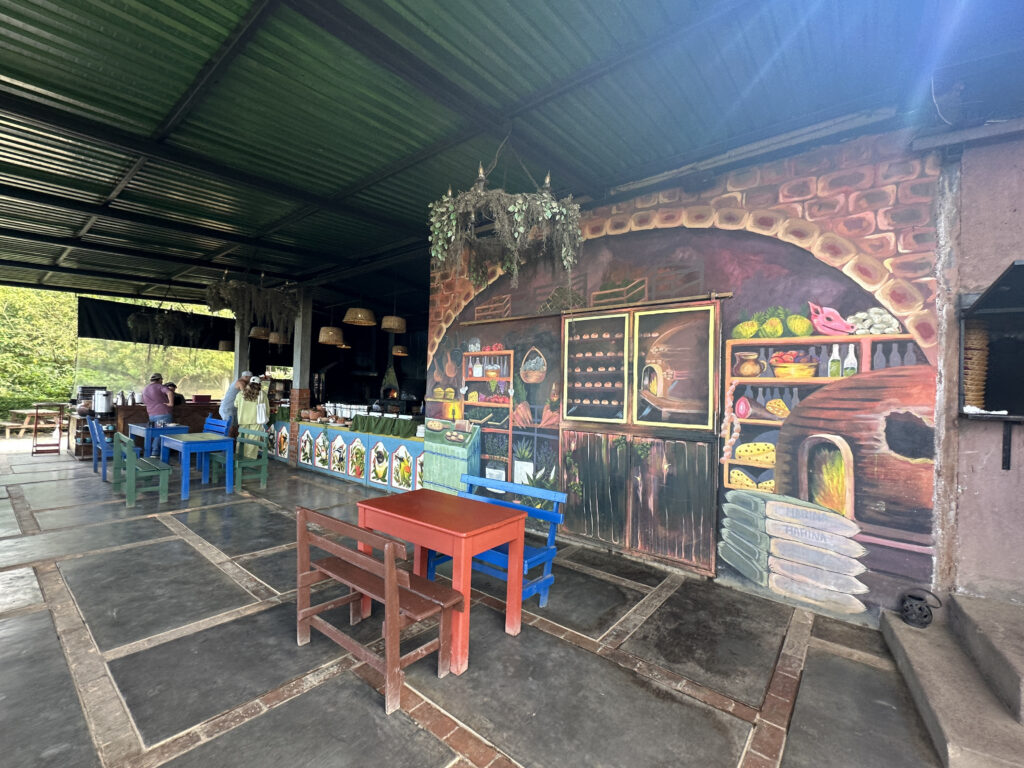 The image shows an outdoor dining area with colorful wooden tables and chairs. The wall features a large, vibrant mural depicting a rustic kitchen scene with an oven, shelves of food, and various kitchen items. The area is covered by a green metal roof, and there are hanging plants and lights. In the background, a couple of people are standing near a counter.