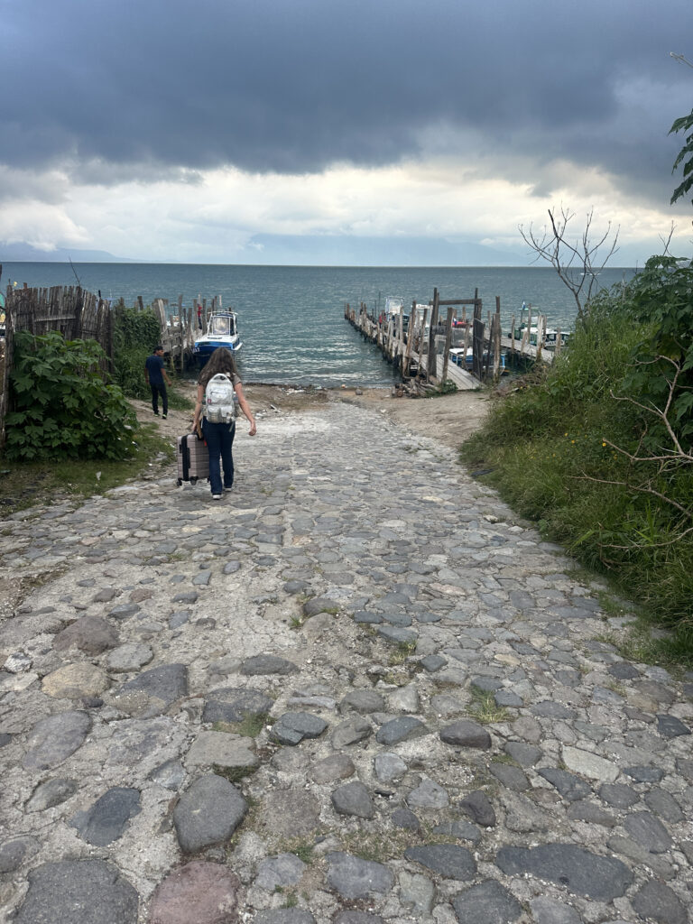 A person with a backpack and a suitcase walks down a cobblestone path towards a wooden dock extending into a large body of water. Another person is walking ahead. The sky is overcast with dark clouds, and there is greenery on either side of the path. Several boats are moored at the dock.