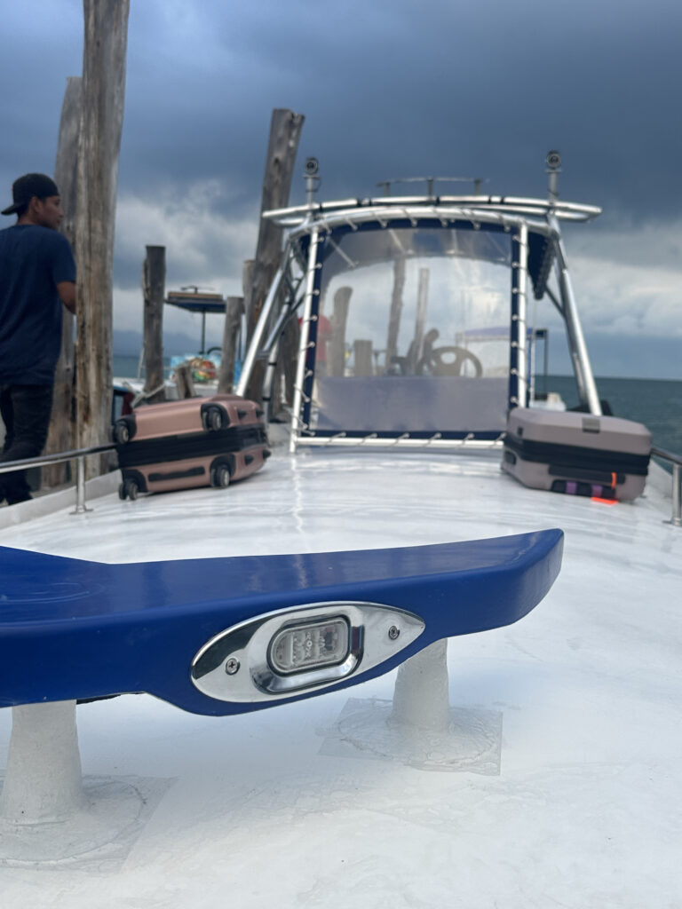 The image shows the deck of a boat with a blue fixture in the foreground. There are several suitcases on the deck, and a person is standing to the left. The background features a canopy structure and a cloudy sky over the ocean.