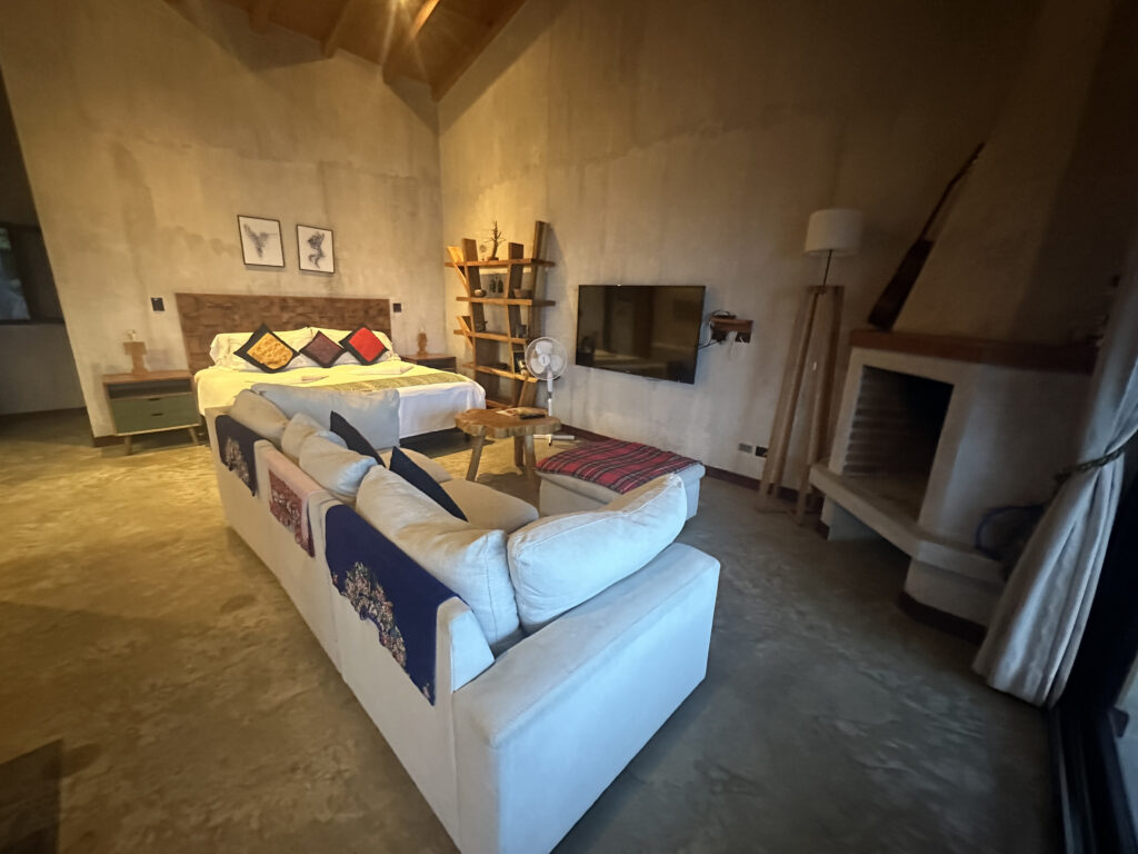 A cozy bedroom with a rustic design. The room features a large bed with colorful pillows and a wooden headboard. In front of the bed is a white sofa with decorative throws. A wooden shelf with various items is against the wall, and a flat-screen TV is mounted nearby. There is a small wooden table and a floor lamp next to a fireplace. The room has a high ceiling and a warm, inviting atmosphere.