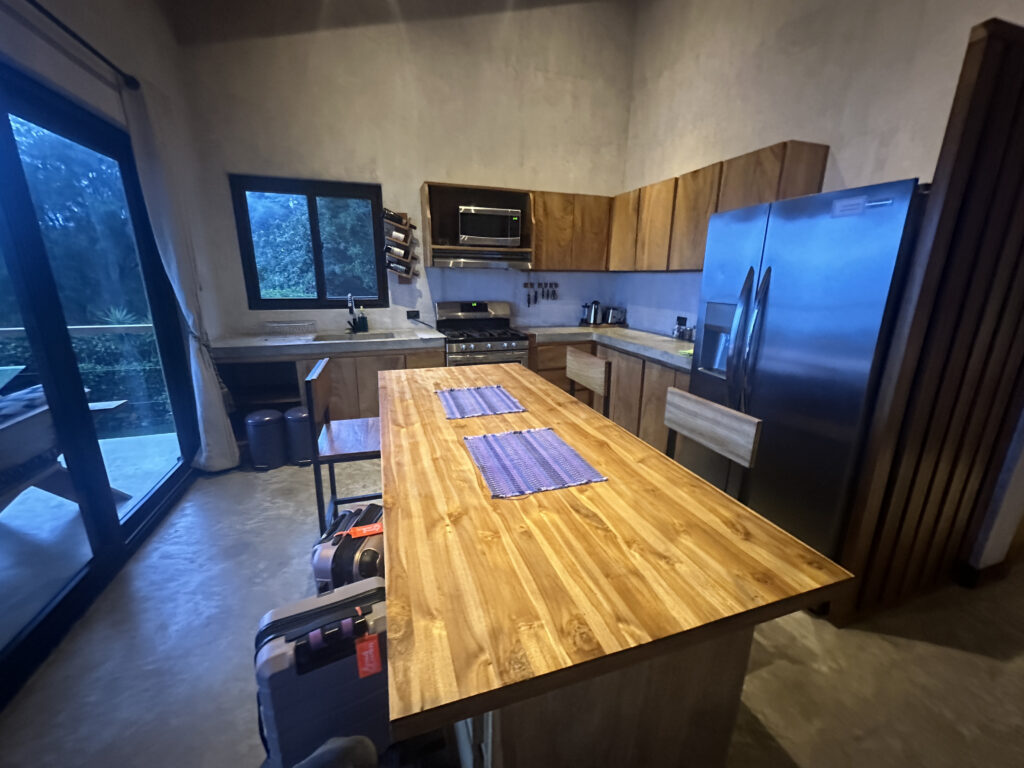 A modern kitchen with wooden cabinets and a large wooden island in the center. The island has two chairs and is set with two blue placemats. Stainless steel appliances, including a refrigerator and a stove, are visible. A window and a sliding glass door provide natural light, and there are suitcases on the floor near the island.