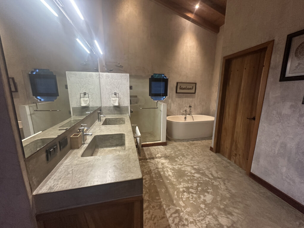 The image shows a modern bathroom with a large mirror and a double sink vanity on the left. The countertop is made of a stone-like material. There are two wall-mounted faucets and a soap dispenser on the counter. A bathtub is visible in the background, positioned against the wall. The walls have a textured finish, and there is a wooden door on the right. The lighting is soft, with ceiling lights providing illumination. A framed picture is hung on the wall near the bathtub.