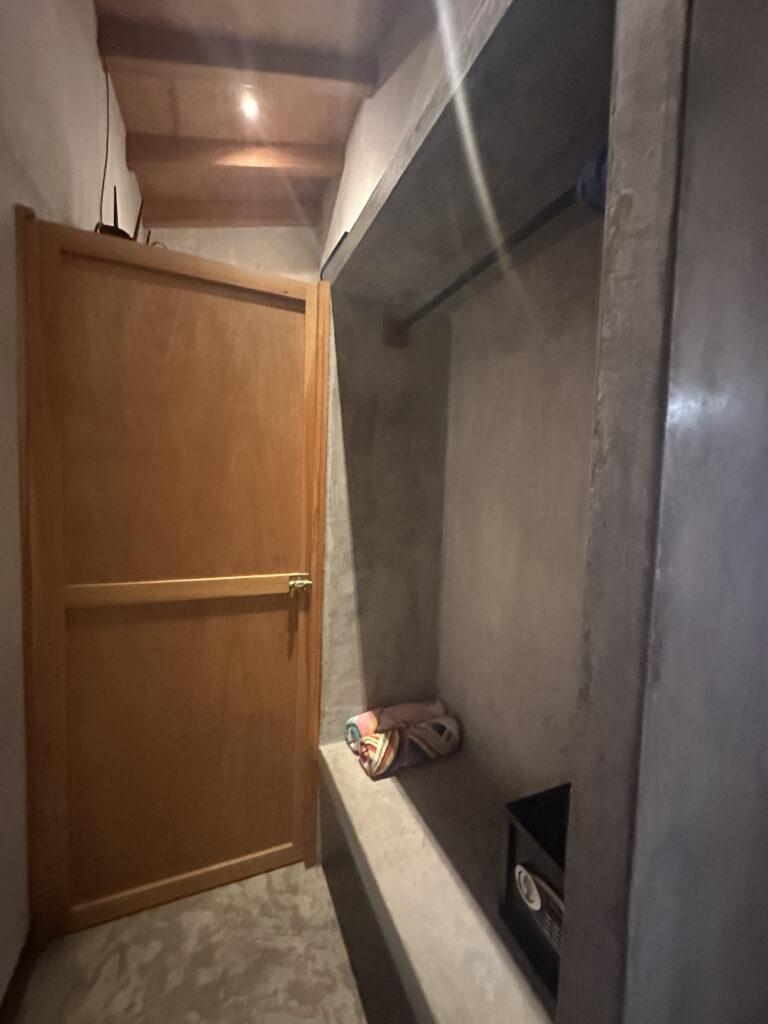The image shows a small, dimly lit room with a wooden door slightly ajar. Inside, there is a concrete shelf with a rolled-up colorful towel and a black box. The ceiling has wooden beams, and a light fixture is visible, casting a warm glow.