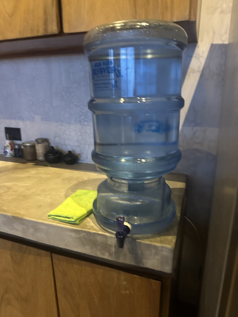 The image shows a large blue water dispenser placed on a kitchen countertop. The dispenser is filled with water and has a spigot at the bottom for dispensing. Next to it, there is a folded green cloth. The countertop is made of a light-colored material, and there are wooden cabinets above and below.