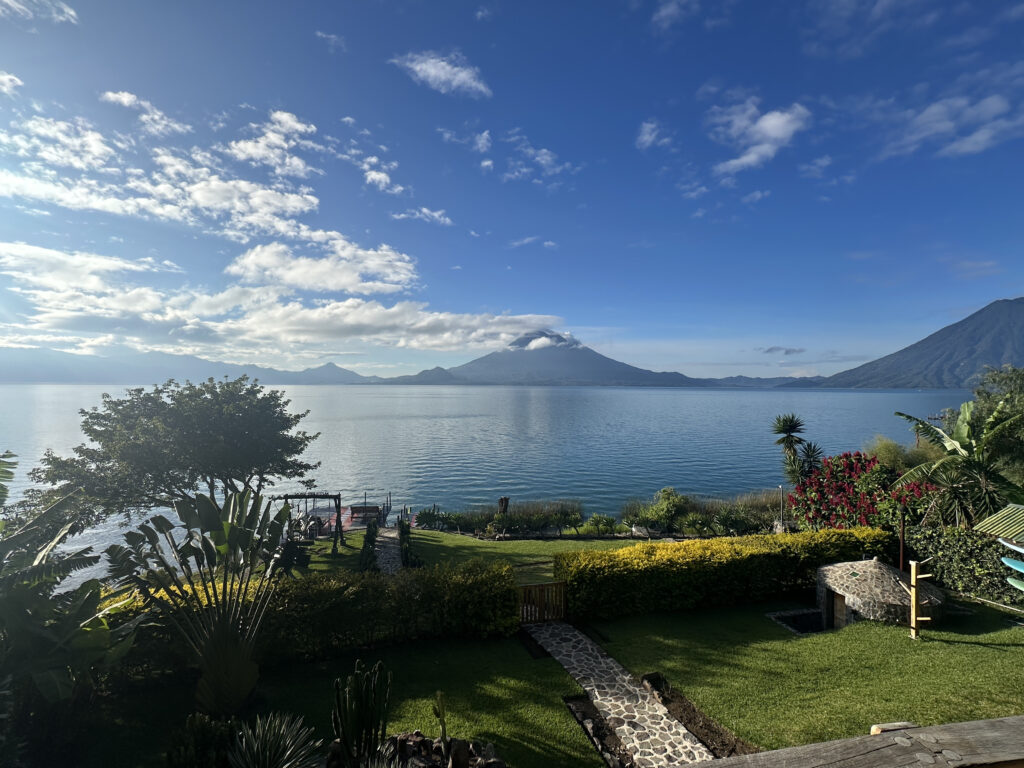 A scenic view of a lake with a mountain in the background under a blue sky with scattered clouds. In the foreground, there is a well-maintained garden with various plants, trees, and a stone pathway leading towards the water. The landscape is lush and vibrant, with a mix of greenery and colorful flowers.