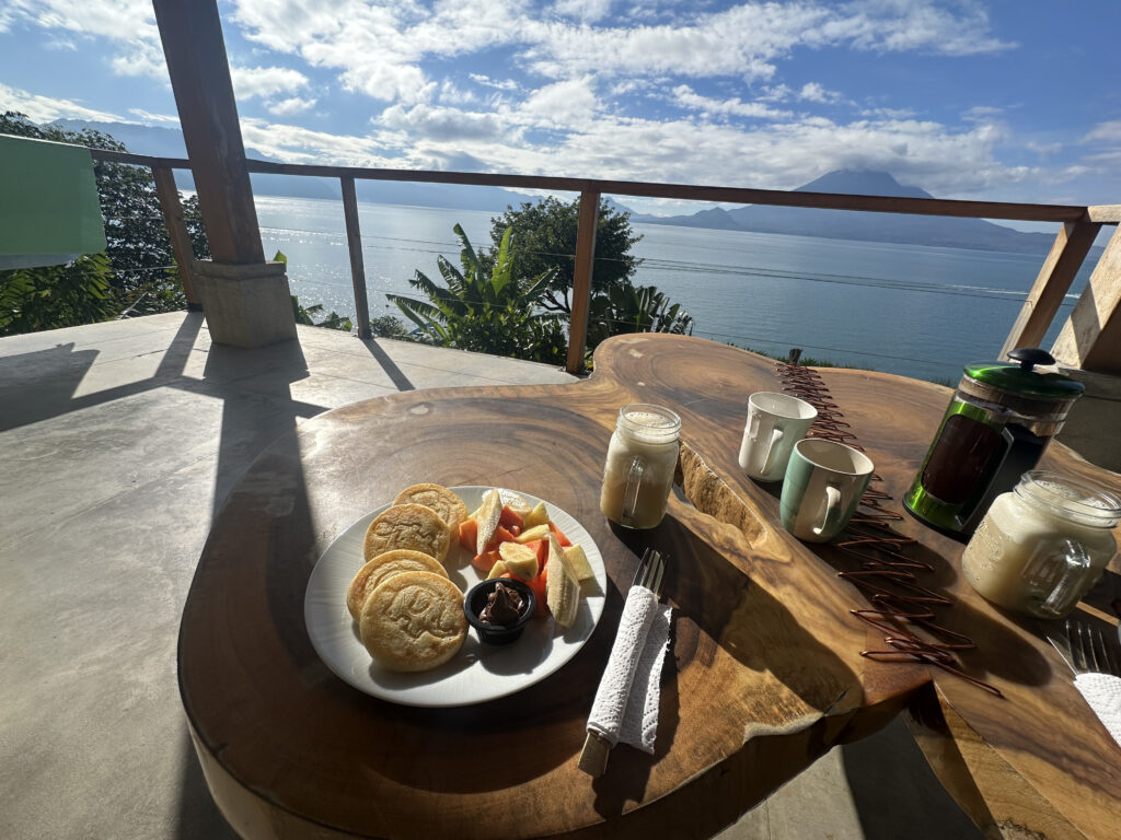 The image shows a wooden table set with a breakfast scene overlooking a scenic view of a lake and mountains. On the table, there is a plate with pancakes and fruit, a small dish of chocolate spread, a French press, two mugs, and a jar of milk. The background features a clear sky with some clouds and lush greenery.