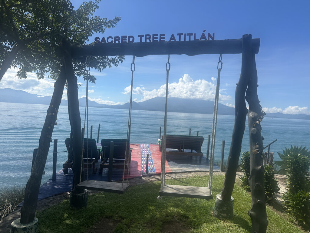 A wooden swing set with two swings is positioned on a grassy area overlooking a large body of water, likely a lake. The top of the swing set has the words "SACRED TREE ATITLÁN" written on it. In the background, there are mountains under a clear blue sky with some clouds. A wooden deck with chairs is visible near the water's edge.