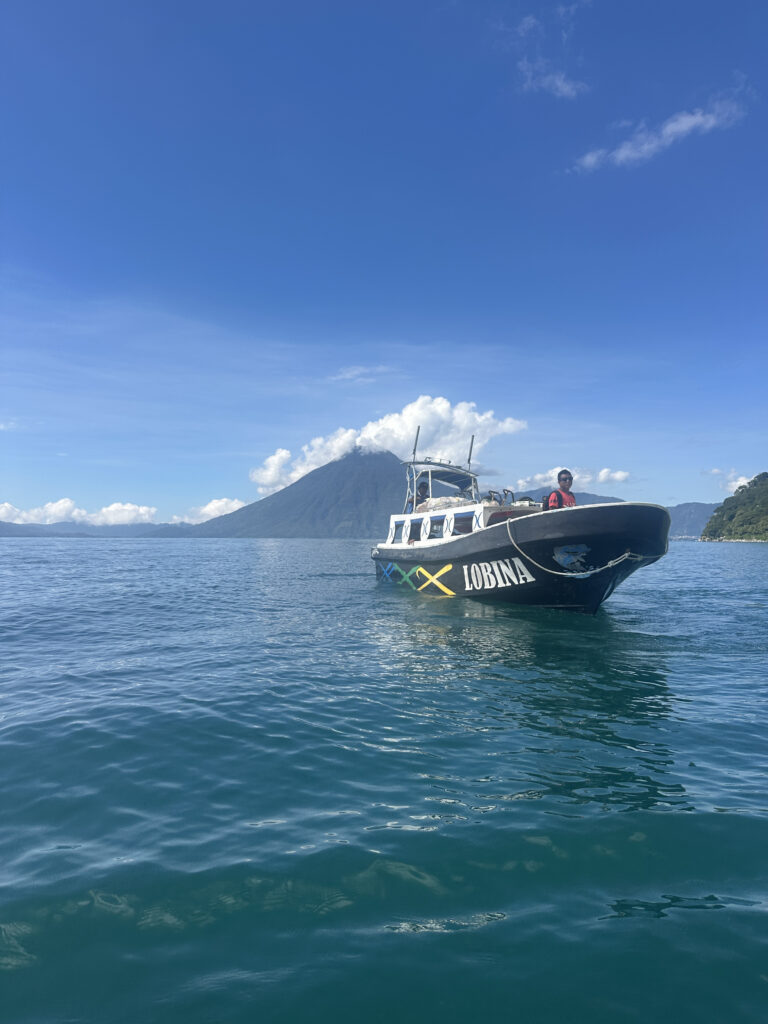 A boat named "Lobina" is floating on a calm body of water. In the background, there is a large mountain with a cloud partially covering its peak. The sky is clear and blue, and a person is visible on the boat.