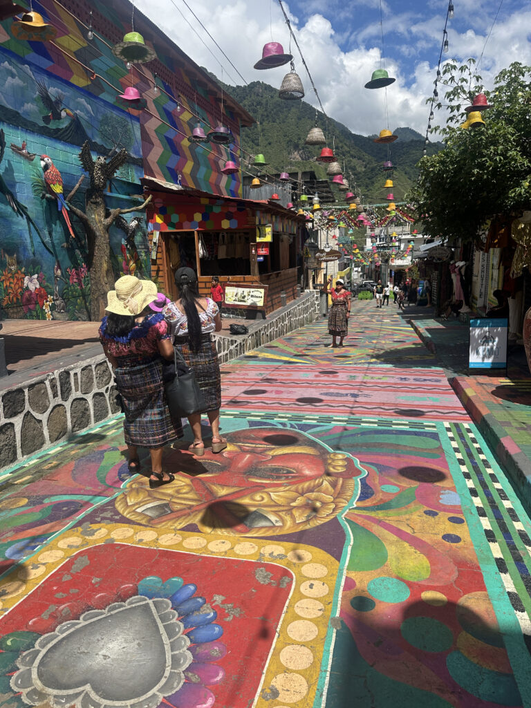 A vibrant street scene in a colorful village. The ground is painted with intricate, multicolored designs, including a large face and heart motif. The walls of the buildings are adorned with murals, including images of birds and geometric patterns. Overhead, strings of colorful hats are suspended, adding to the festive atmosphere. People in traditional clothing are walking along the street, and lush green mountains are visible in the background under a partly cloudy sky.