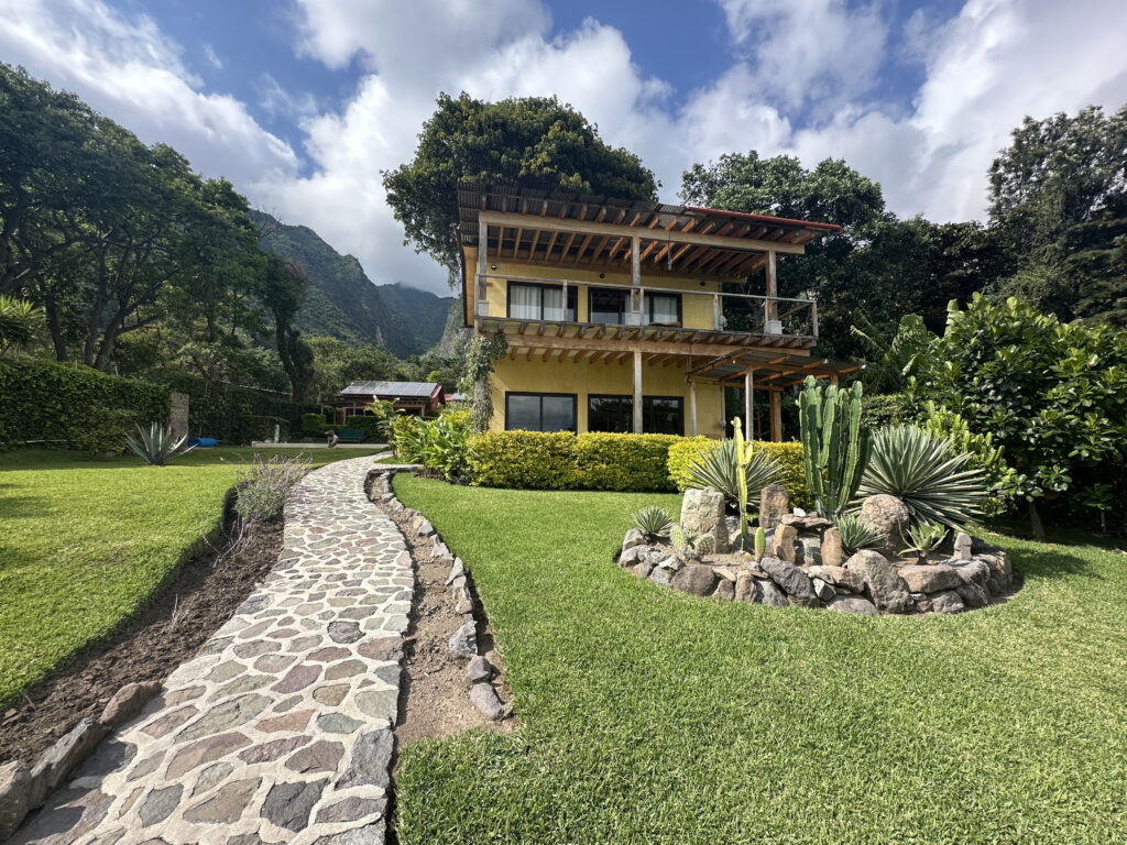 A two-story yellow house with a wooden balcony is surrounded by lush greenery and trees. In the foreground, there is a stone pathway leading to the house, and a circular rock garden with various cacti and succulents. The background features mountains under a partly cloudy sky.
