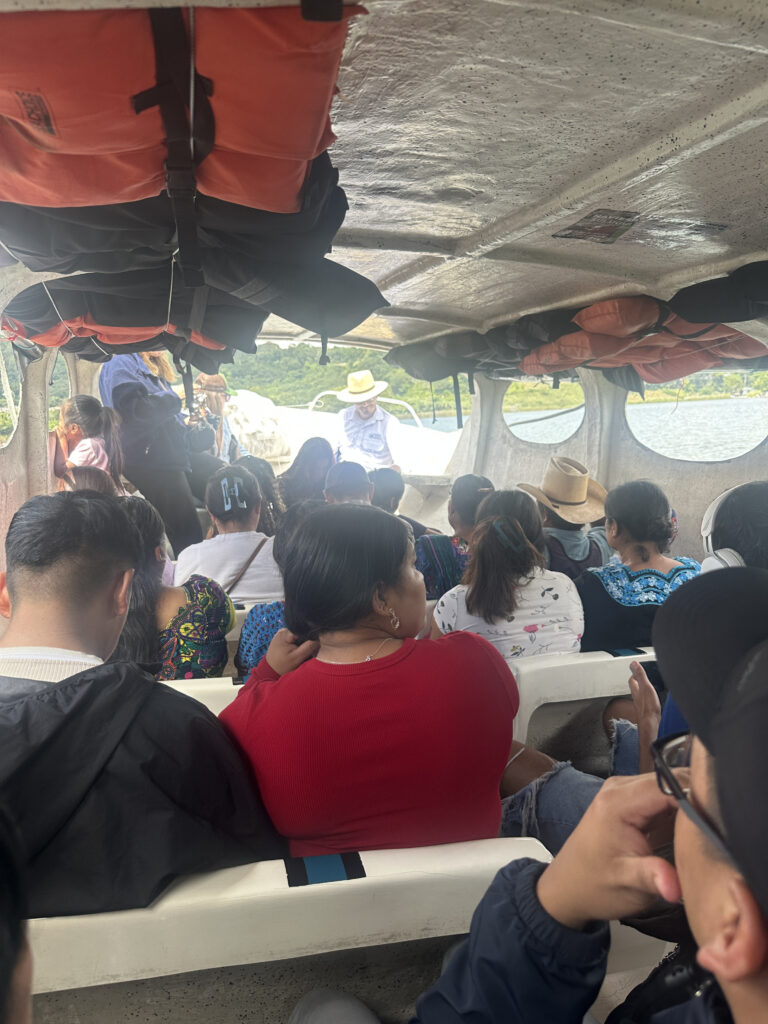 A group of people is seated inside a boat, with life jackets stored above them. The boat is on a body of water, and some passengers are looking out at the scenery. The interior is crowded, and the passengers are wearing casual clothing, including hats and jackets.