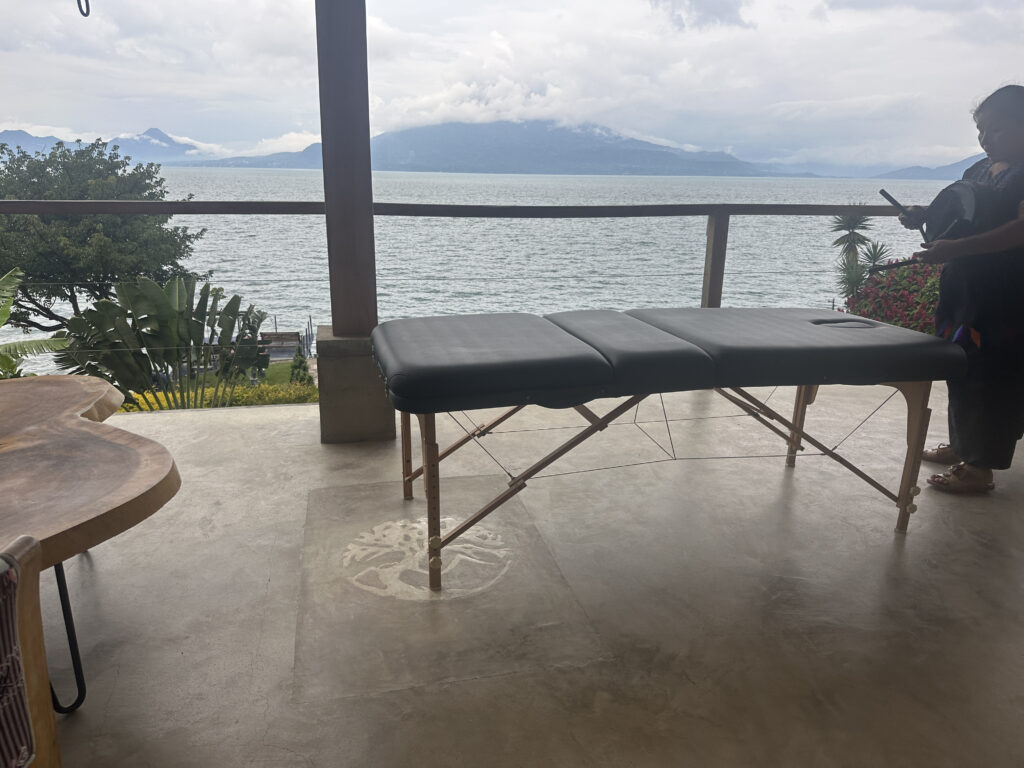 A massage table is set up on a covered patio overlooking a large body of water with mountains in the background. There are trees and plants around the patio, and a person is standing to the right, partially visible, holding an object. The sky is cloudy, creating a serene and tranquil atmosphere.