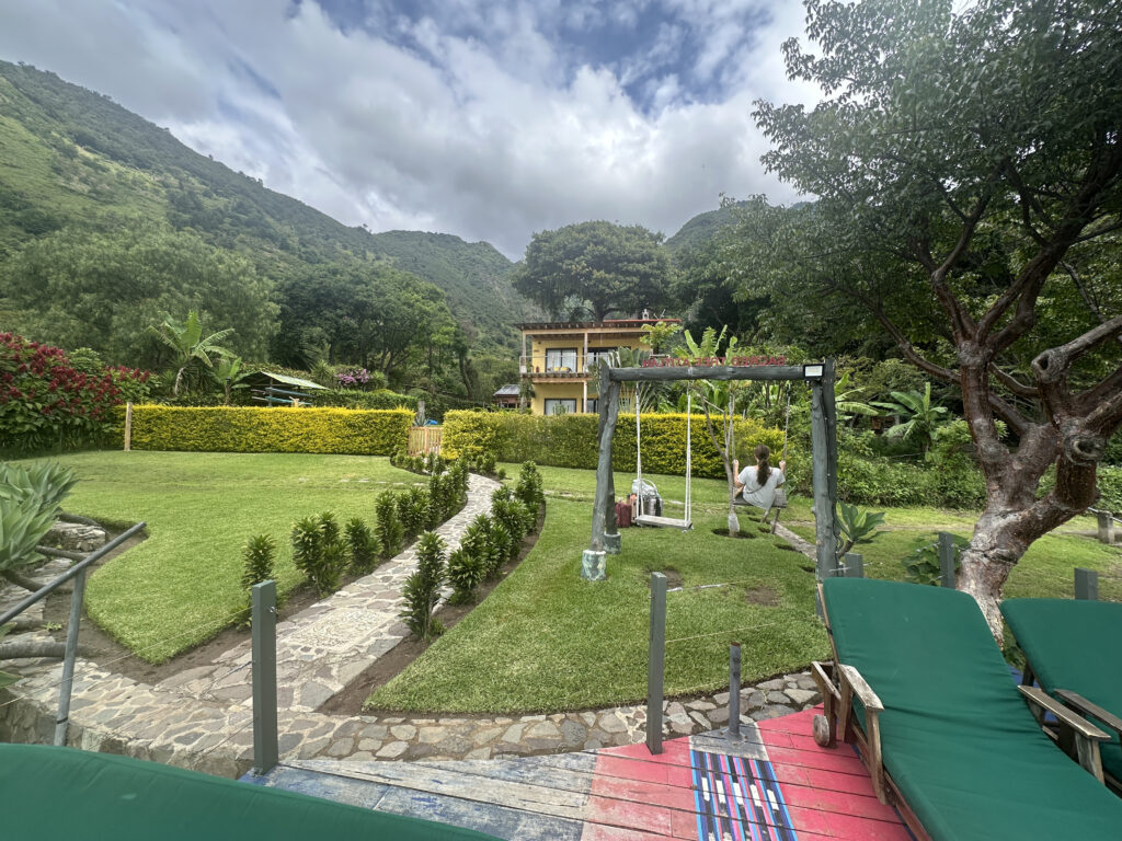 A scenic garden with a stone path leading through well-manicured grass and small shrubs. A person is sitting on a swing under a wooden frame, surrounded by lush greenery and trees. In the background, there is a two-story house with a mountainous landscape under a cloudy sky. Two green lounge chairs are in the foreground.