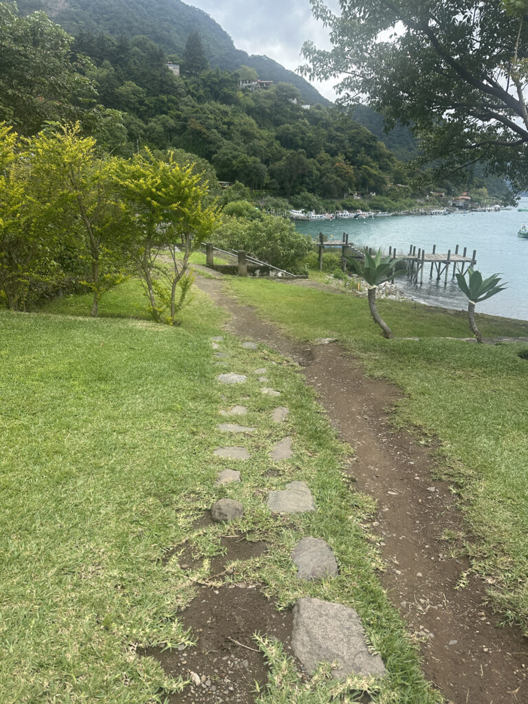 A scenic view of a grassy path leading down to a wooden dock by a lake. The path is lined with stones and surrounded by lush greenery and trees. In the background, there are forested hills and a cloudy sky. The lake is calm, with several boats docked along the shoreline.