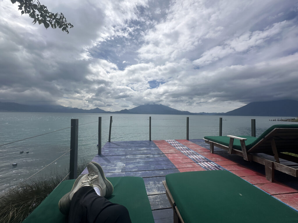 A scenic view of a lake with mountains in the background under a cloudy sky. In the foreground, a person is relaxing on a green cushioned lounge chair on a deck, with their legs visible. Another empty lounge chair is nearby. The deck is painted in red and blue stripes, and there is a railing along the edge.