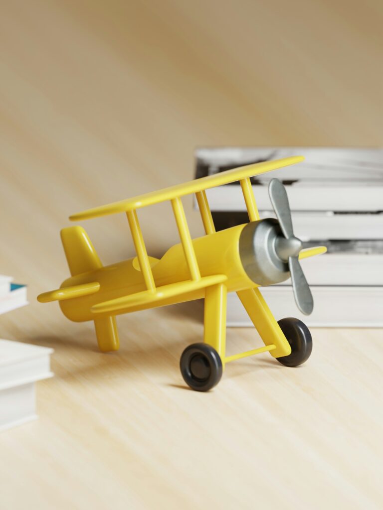 A yellow toy biplane with a silver propeller is placed on a light wooden surface. In the background, there are stacks of books with white and black covers. The scene is softly lit, creating a warm and playful atmosphere.