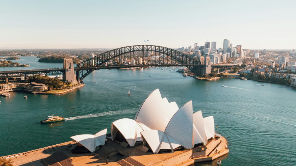 Aerial view of Sydney Harbour, featuring the Sydney Opera House in the foreground with its distinctive sail-like design. The Sydney Harbour Bridge spans the water in the background, connecting the cityscape on both sides. The water is dotted with boats, and the skyline is visible under a clear blue sky.
