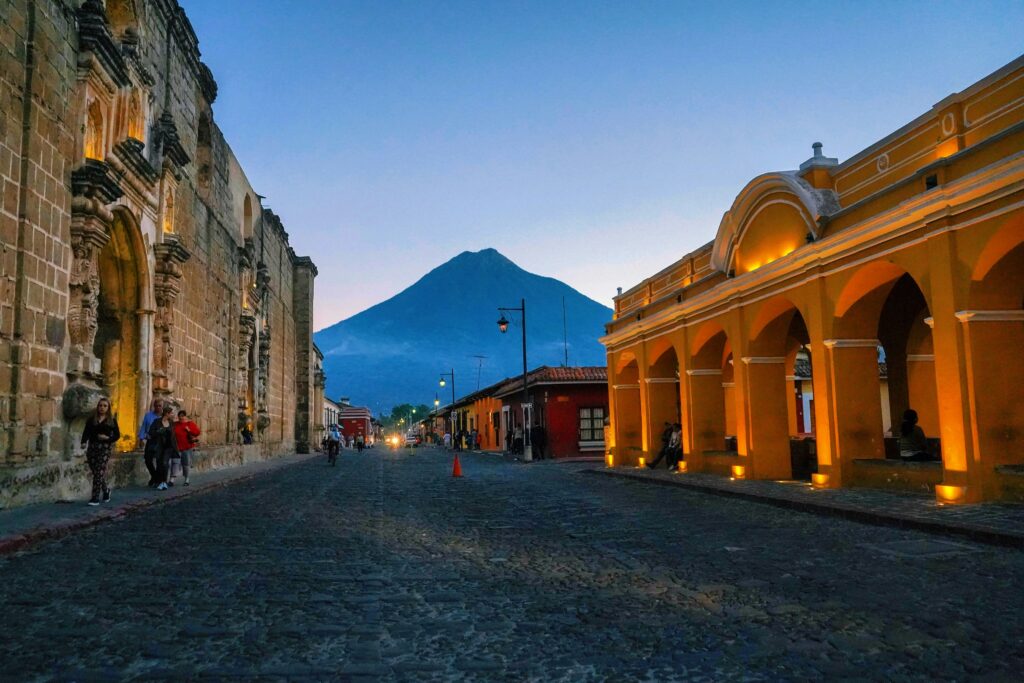 A cobblestone street in a historic town at dusk, with people walking and a cyclist. On the left, there is an old stone building with intricate carvings, and on the right, a yellow building with arches lit by warm lights. In the background, a large volcano is visible against the evening sky.