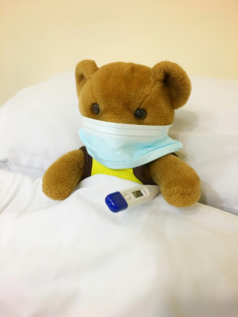 A teddy bear is sitting on a bed, covered with a white blanket. It is wearing a light blue surgical mask and has a digital thermometer tucked under its arm.
