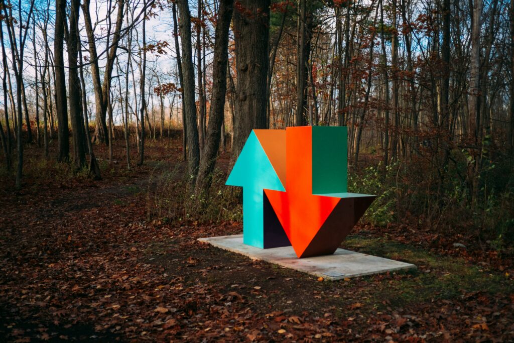 A colorful sculpture in the shape of an upward and downward arrow is placed on a concrete slab in a forest. The sculpture is painted in bright shades of teal, orange, and red. The surrounding area is filled with tall trees and fallen leaves, suggesting an autumn setting.