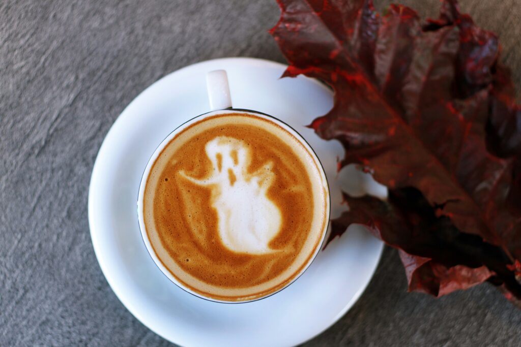 A cup of coffee with latte art depicting a ghost on the surface, placed on a white saucer. Next to the cup, there is a large, dark red autumn leaf. The background is a textured gray surface.