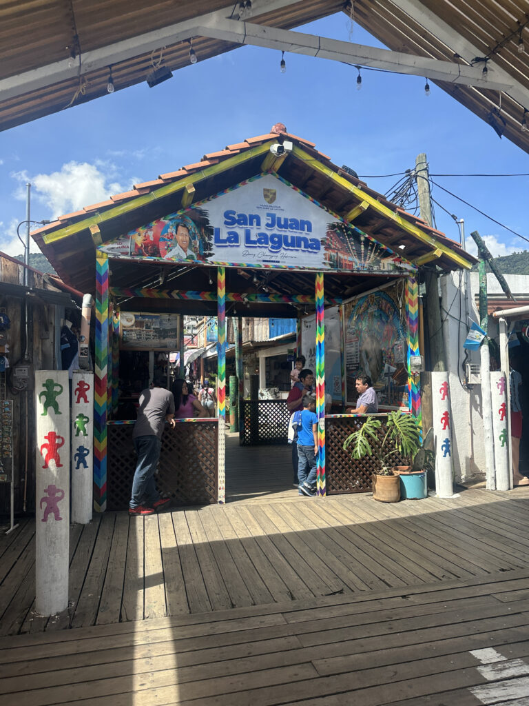 The image shows a colorful entrance to a building or market area in San Juan La Laguna. The structure has a wooden roof with a sign displaying the name "San Juan La Laguna." The pillars and beams are decorated with vibrant, multicolored patterns. There are people standing and interacting near the entrance, and the ground is made of wooden planks. The sky is clear and blue, suggesting a sunny day.