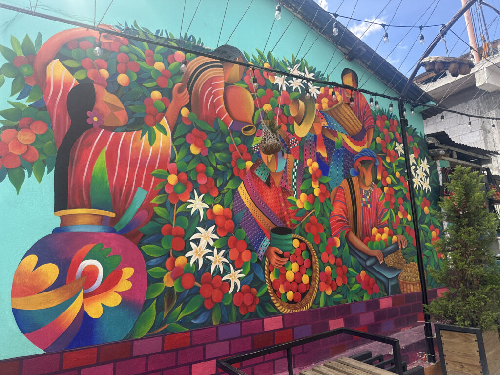 A vibrant mural depicting a colorful scene of people engaged in agricultural activities. The artwork features individuals in traditional clothing, surrounded by lush greenery and flowers. They are holding baskets filled with fruit, and one person is pouring from a large jug. The mural is painted on the side of a building, with string lights hanging above and a small tree in a planter nearby.