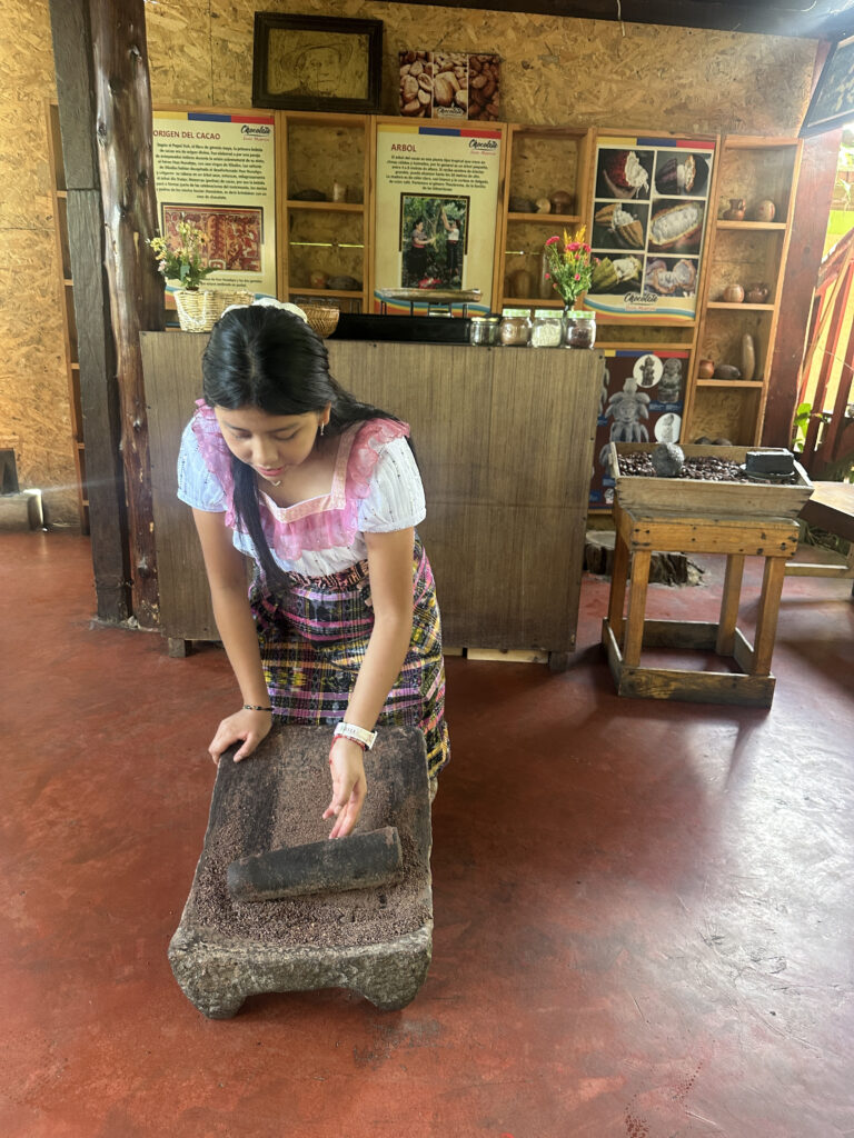 A woman is using a traditional stone tool to grind cocoa beans. She is wearing a colorful, traditional outfit and is focused on her task. Behind her, there are informational posters about cocoa and various items on display, including jars and flowers. The setting appears to be an educational or cultural exhibit related to cocoa production.