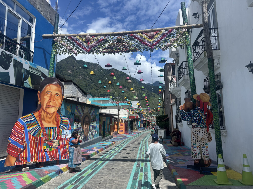 A vibrant street scene features colorful murals and decorations. On the left, a large mural of an elderly woman in traditional clothing is painted on a blue wall. The street is adorned with colorful patterns and overhead decorations, including hanging flowers and multicolored hats. People are walking along the street, and the background shows green mountains under a partly cloudy sky.