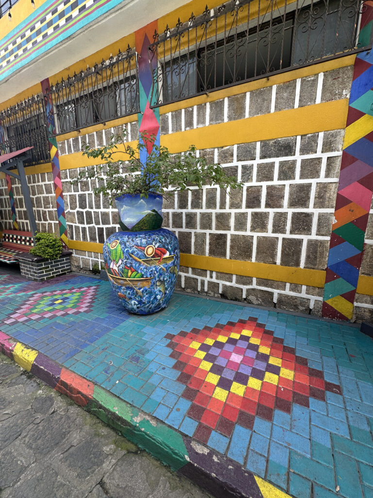 The image shows a vibrant, colorful street scene. A large, decorative ceramic pot with a plant is placed on a sidewalk. The pot features intricate designs, including a landscape and abstract patterns. The sidewalk is made of blue tiles with colorful diamond patterns. The building wall behind the pot is painted with a mix of yellow, white, and multicolored geometric designs, and has barred windows. The overall scene is lively and artistic.