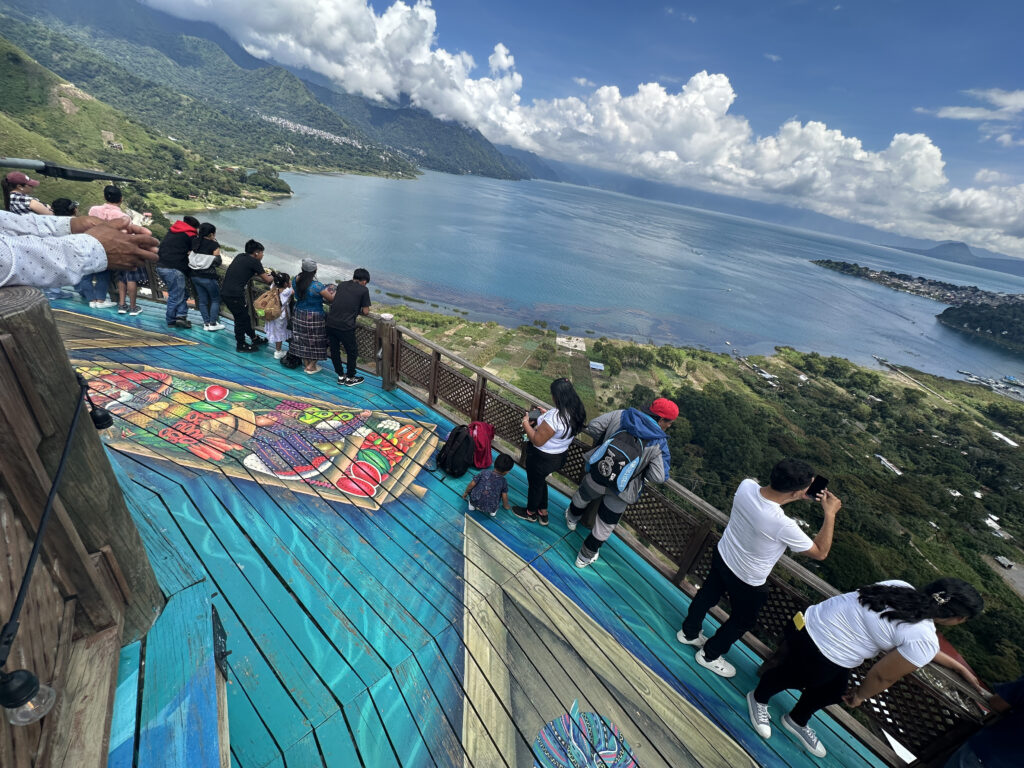A group of people is standing on a colorful wooden platform with a vibrant mural, overlooking a scenic view of a large lake surrounded by lush green hills and mountains. The sky is partly cloudy, and the water is a deep blue. Some people are taking photos, while others are enjoying the view.