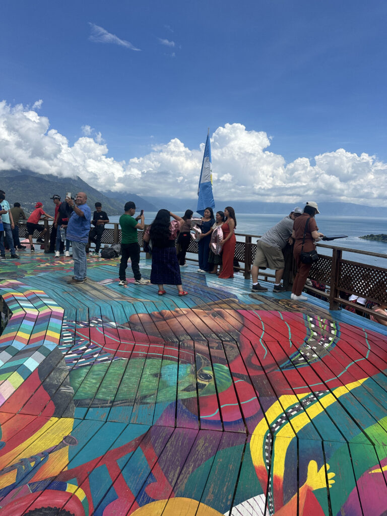 A group of people is standing on a colorful wooden deck with a large mural painted on it. The mural features vibrant, abstract designs and a face. In the background, there is a scenic view of a lake and mountains under a blue sky with clouds. A flag is visible near the group of people.